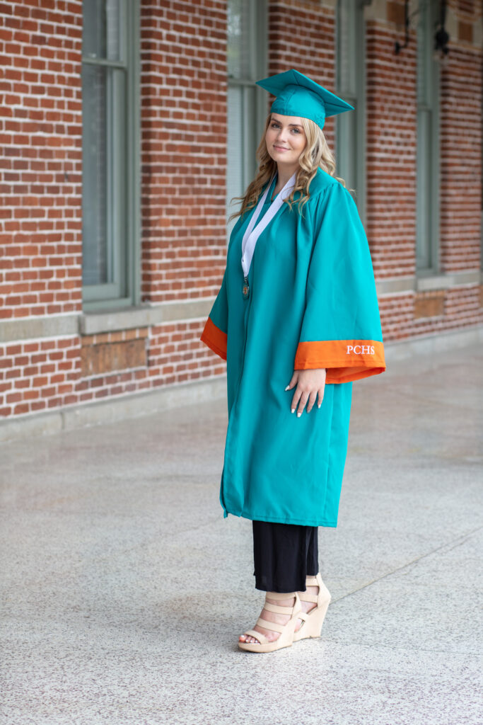 graduation girl in her cap and gown standing on a patio with brick wall in background
