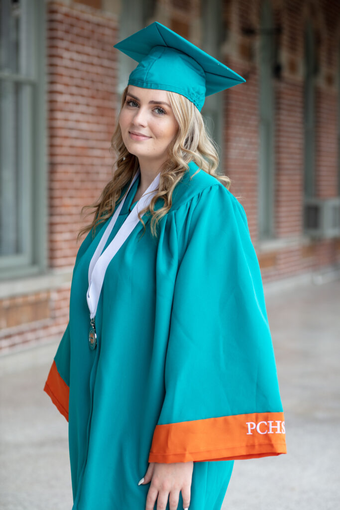 graduation girl in her cap and gown standing on a patio with brick wall in background