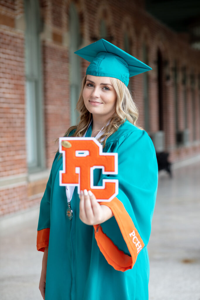 graduation girl in her cap and gown standing on a patio with brick wall in background