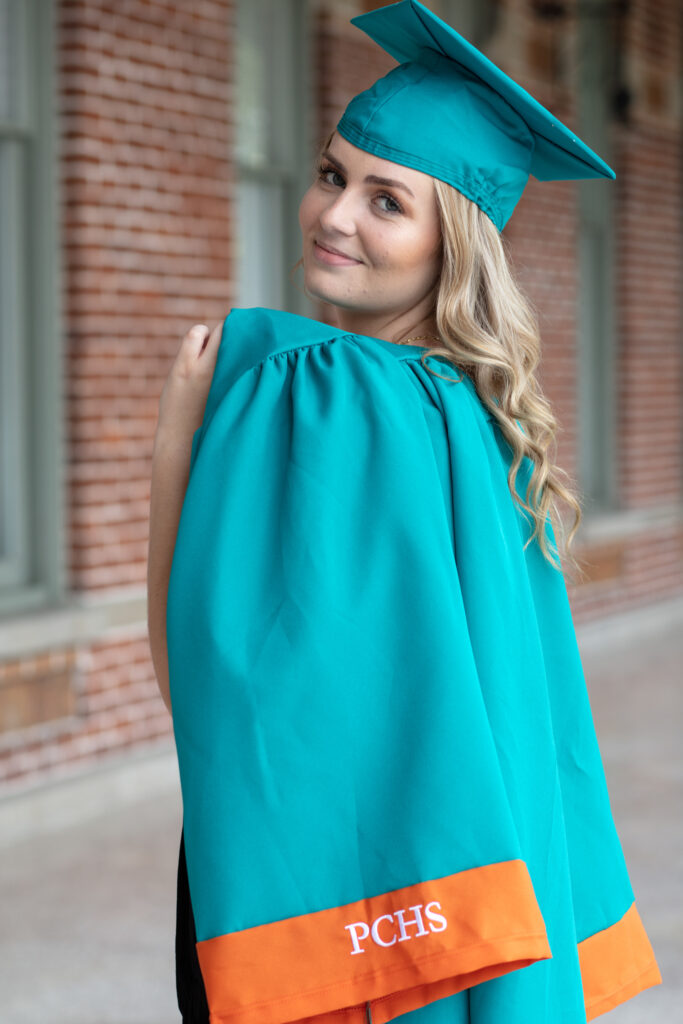 graduation girl in her cap and gown standing on a patio with brick wall in background
