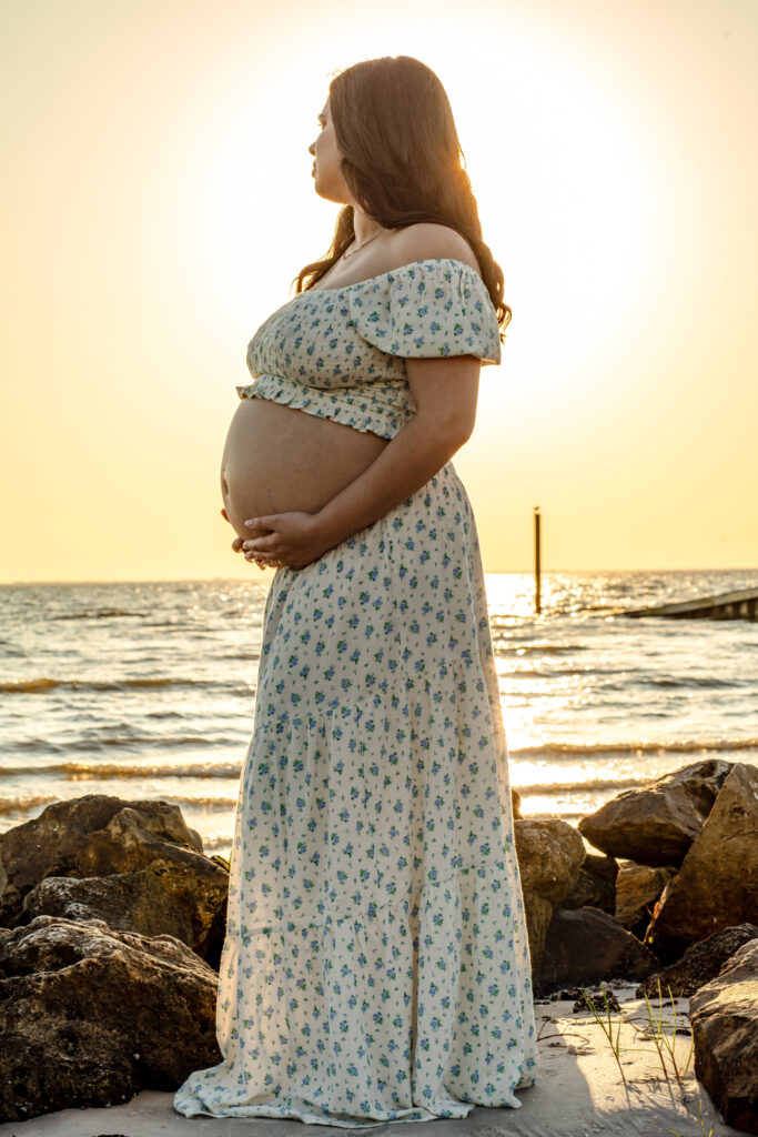momma holding her belly standing on the rocks in a rustic dress at sunset