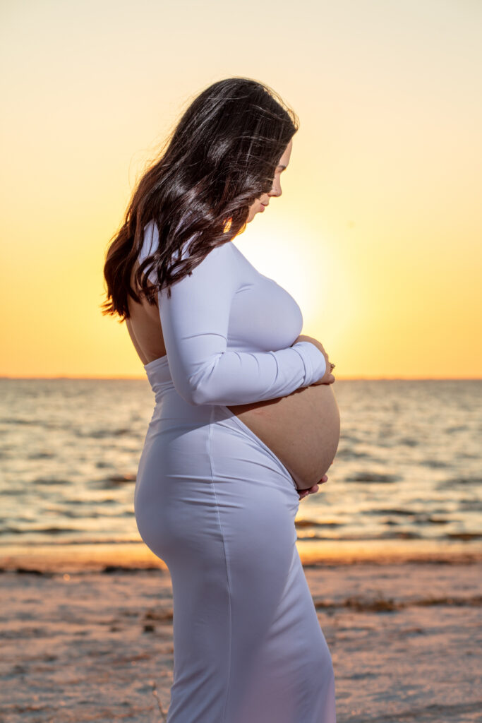 momma holding her pregnant belly in a white dress on the beach at sunset