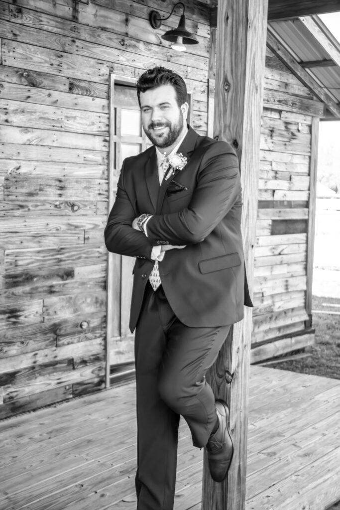 Groom standing against a wooden barn pole with his foot up against the pole