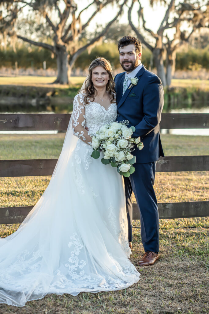 Bride and groom posing for the camera in front of a wooden fence on a farm