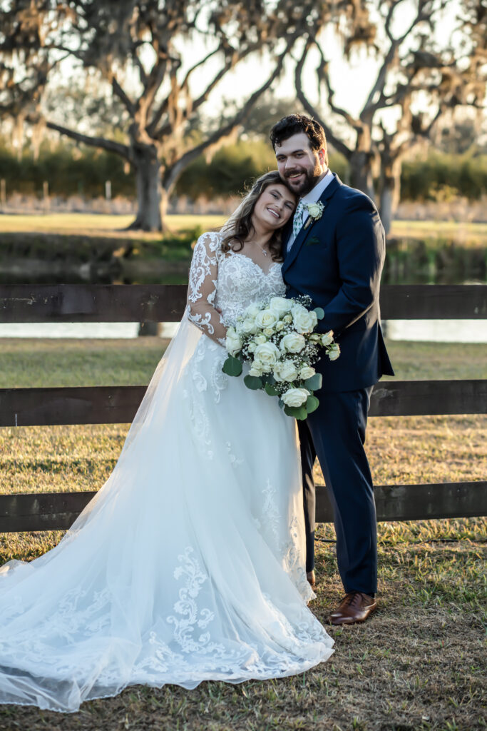 Bride leaning on the Groom's shoulder standing in front of a wooden fence