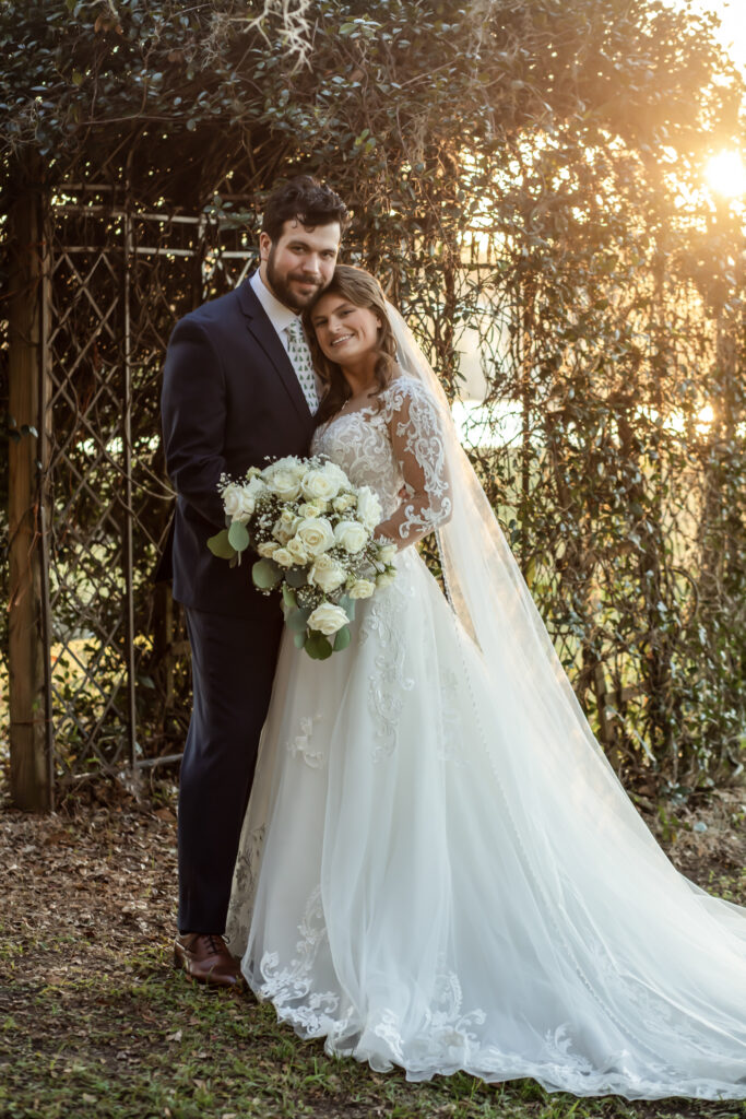 Bride leaning on the groom's shoulder with leafy lattice and the sunset behind them