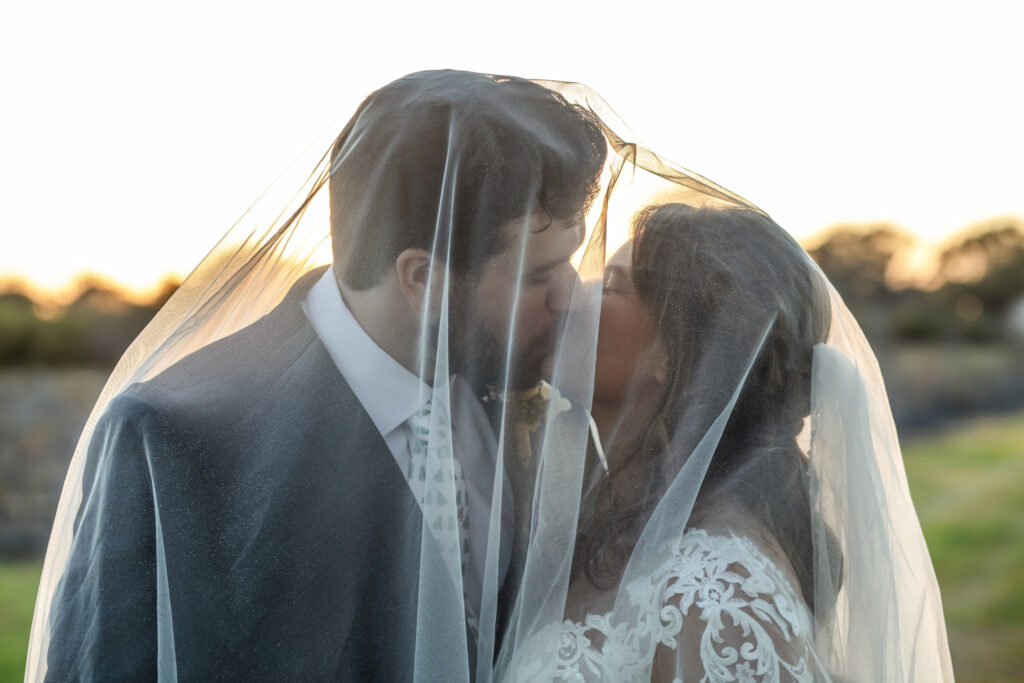 Bride and groom kissing under the brides veil with the sunset in the background