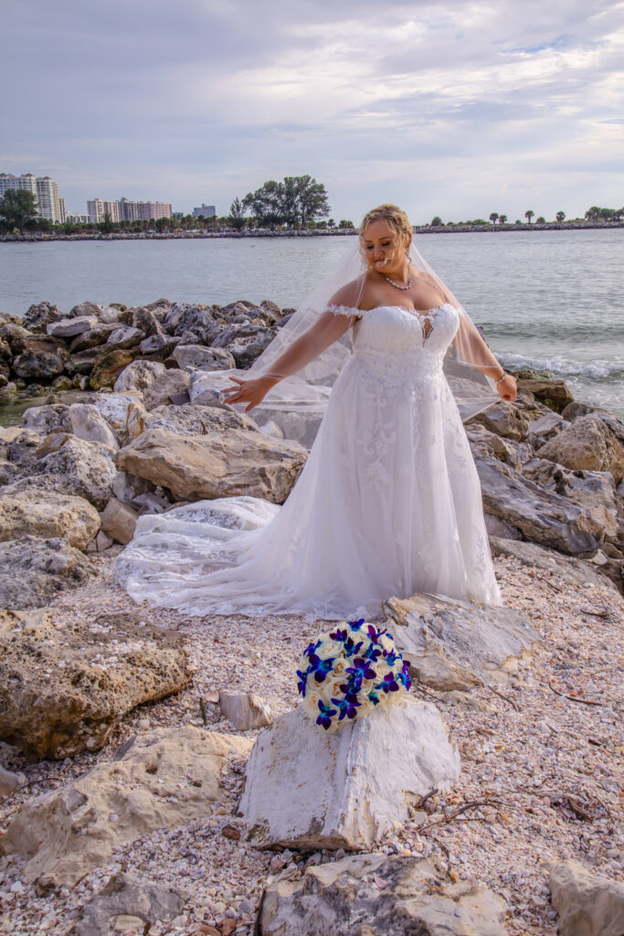 bride in her wedding dress, spreading her veil out with her arms, standing on some rocks on the beach