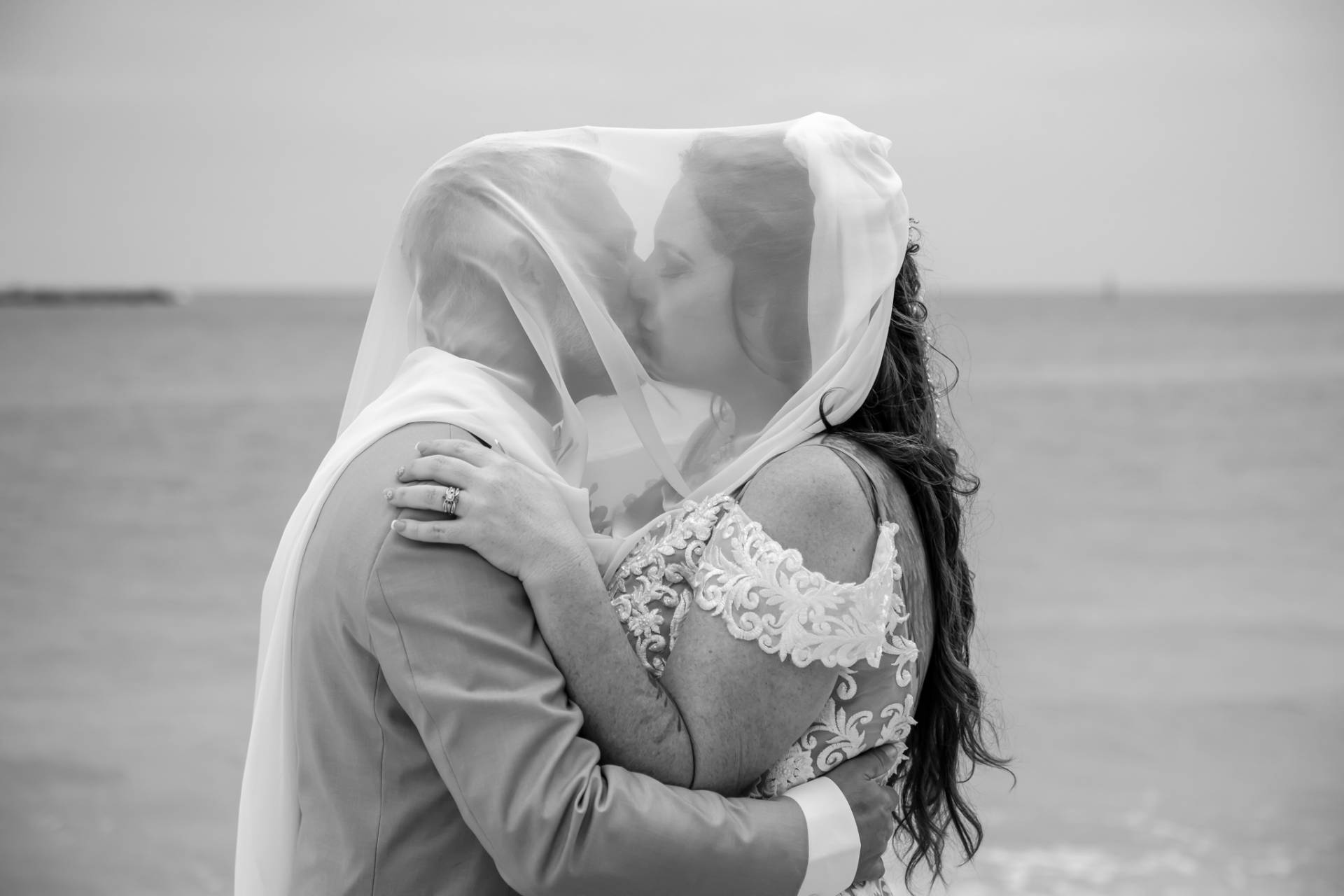 brides veil over her and the groom's heads, on the beach