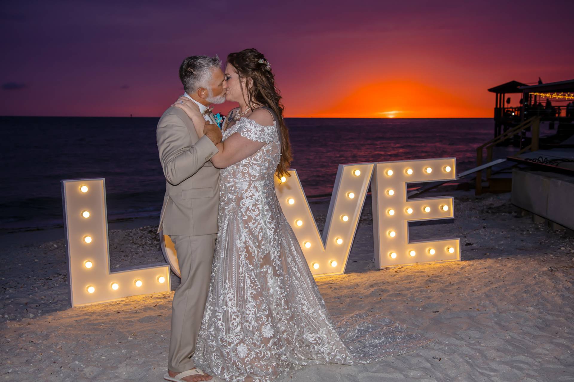 bride and groom standing in front of love LED lights, at sunset, on the beach
