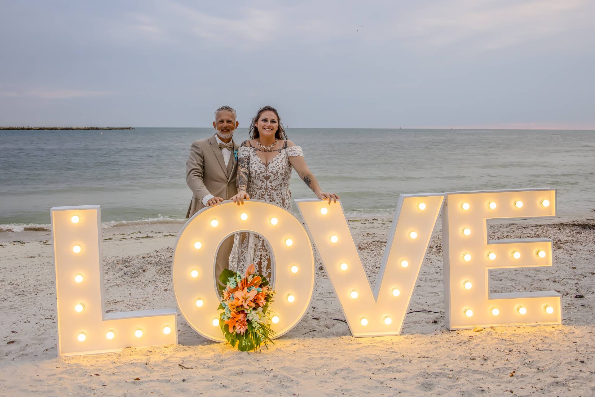 bride and groom standing behind a love LED sign on the beach at sunset
