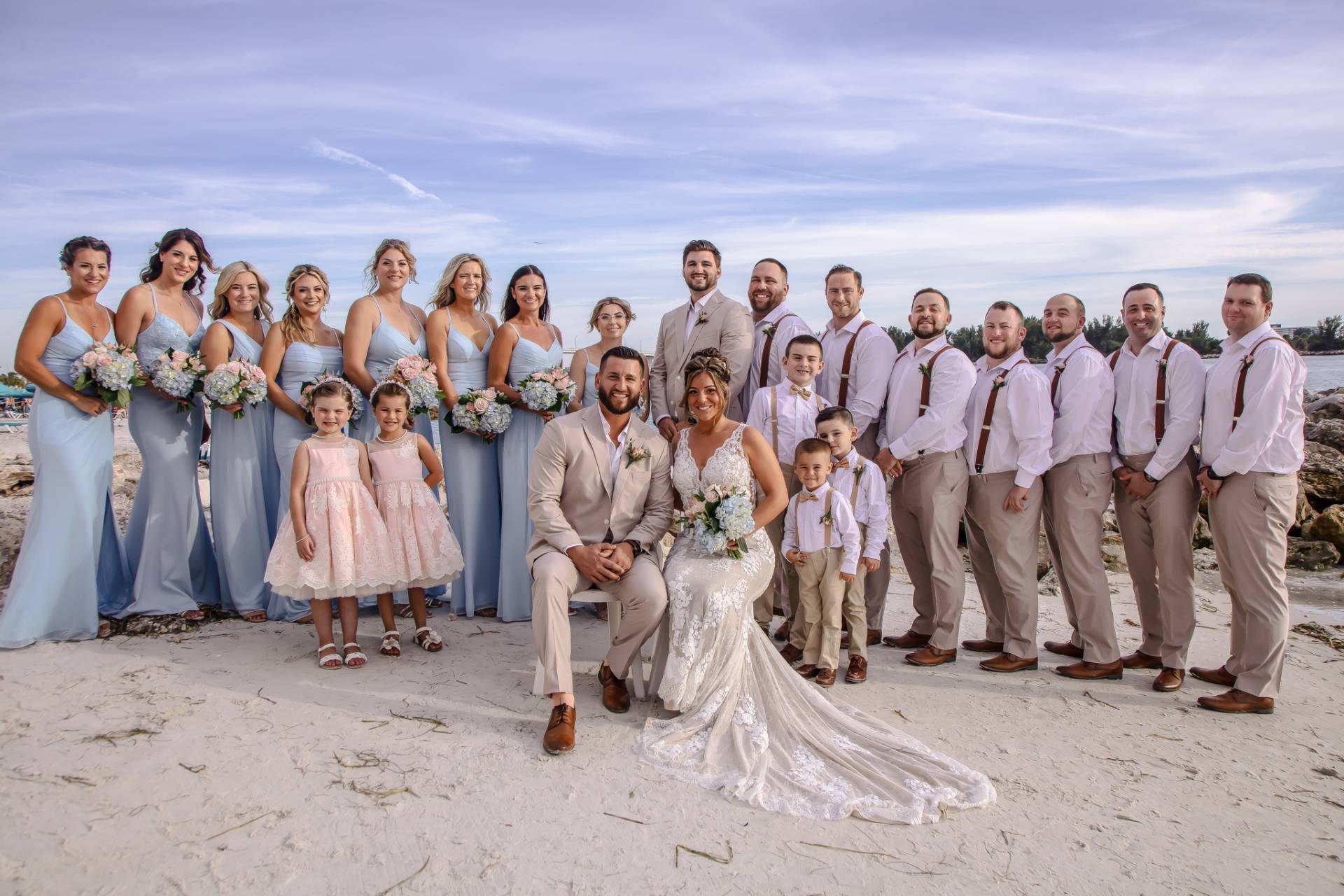 bride and groom sitting in chairs with bridal party standing behind them on the beach